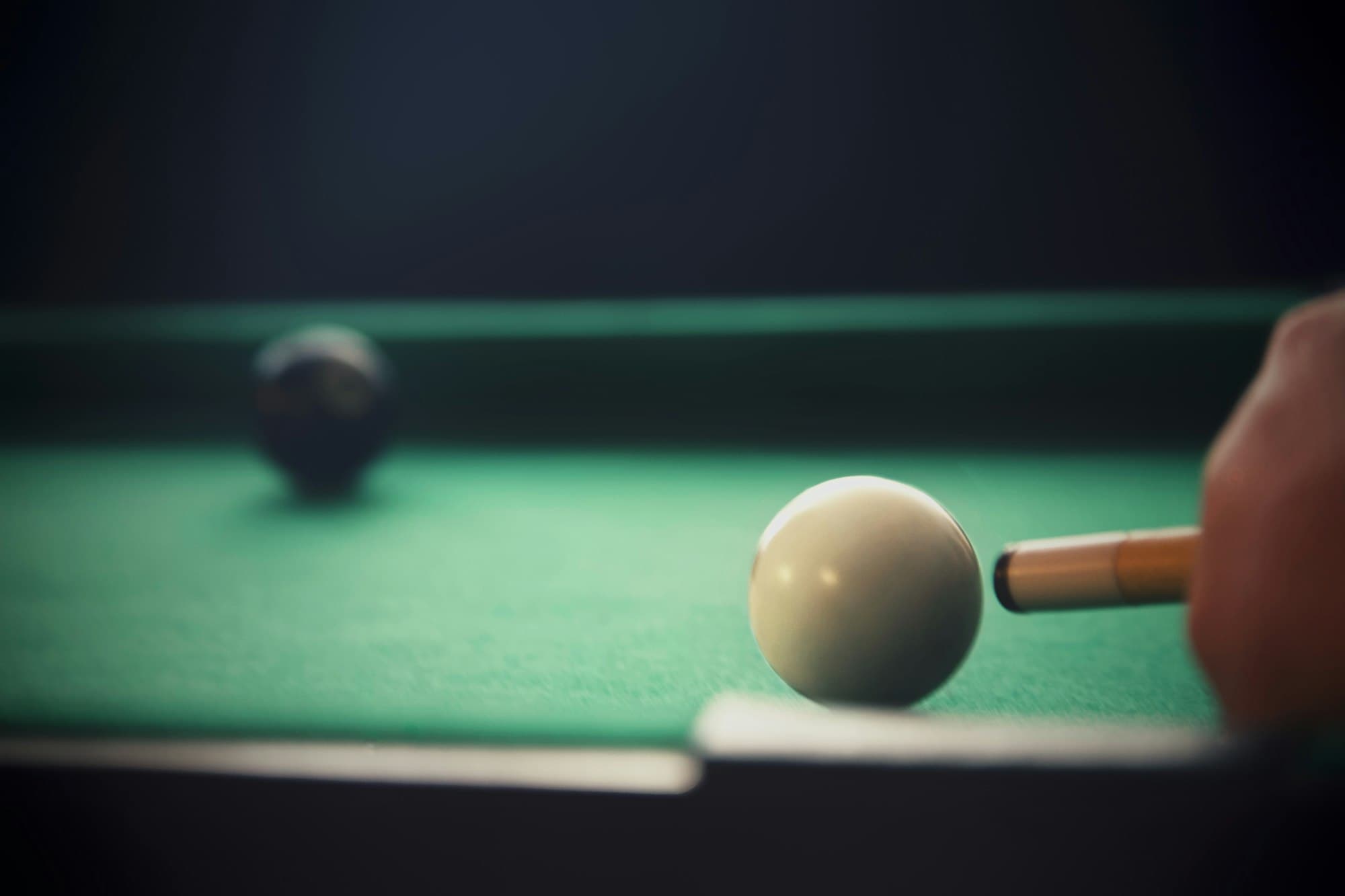 Ujung stik hampir menyentuh bola putih di atas meja berkain hijau, satu bola hitam buram di belakangnya